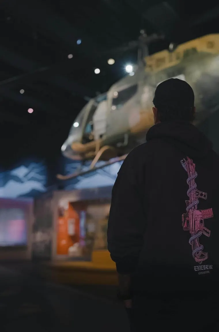 Heavy hooded operator in a dark hangar, rifle-and-serpent print down the back.