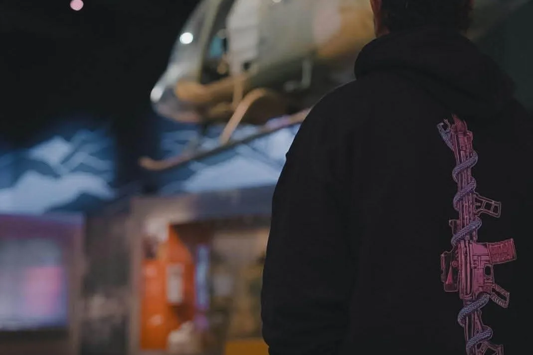 Heavy hooded operator in a dark hangar, rifle-and-serpent print down the sleeve.